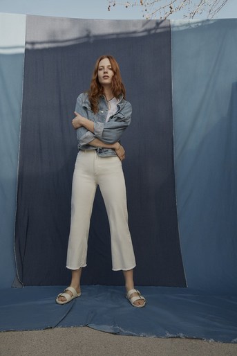 A young woman poses in a denim jacket and white pants against a blue backdrop, striking a casual pose with arms crossed in a studio setting.