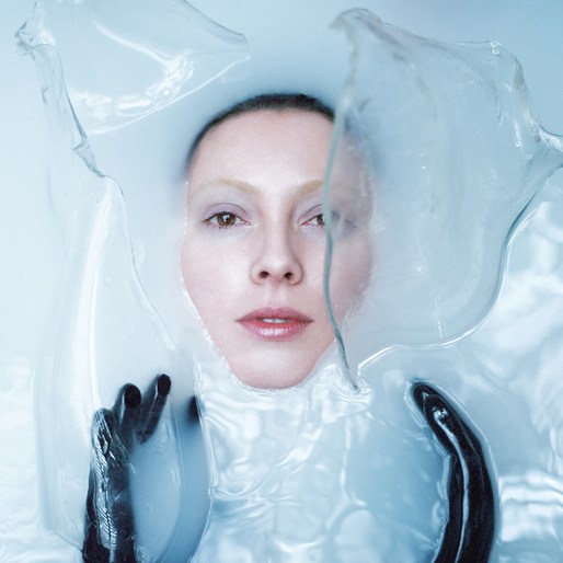 Woman in liquid, face and shoulders above the surface, wearing black gloves, surrounded by clear shapes, pale skin, and neutral expression.
