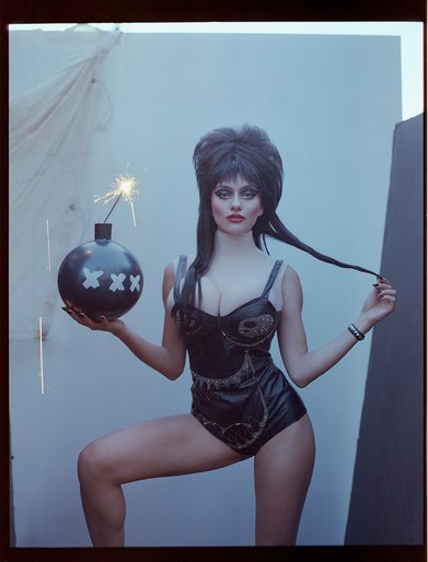 Portrait of a woman dressed as Elvira holding a black cartoon bomb with a sparkler, against a studio backdrop, fashion photography.