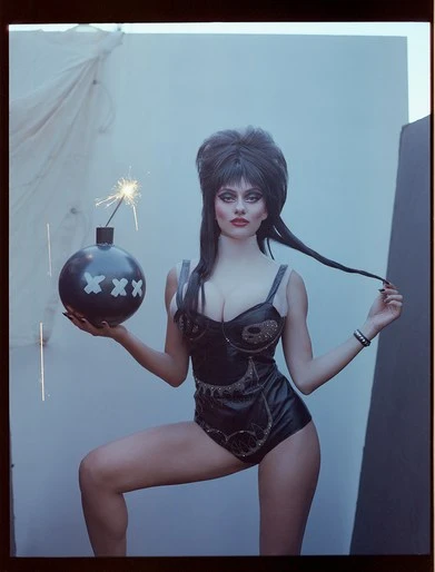 Portrait of a woman dressed as Elvira holding a black cartoon bomb with a sparkler, against a studio backdrop, fashion photography.