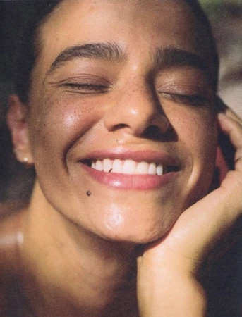 Close-up portrait of a smiling woman with freckles and eyes closed, resting her chin on her hand, by Bryn Bowen.