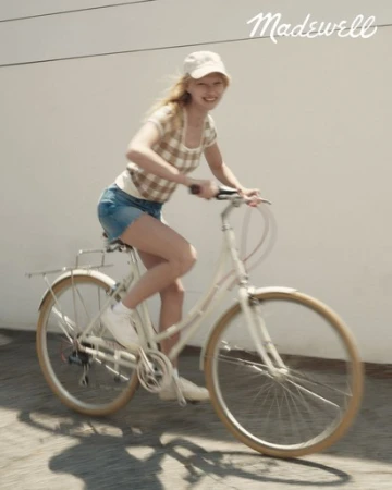 A smiling young woman rides a white bicycle down a concrete path, wearing a patterned top, denim shorts, and a white baseball cap.