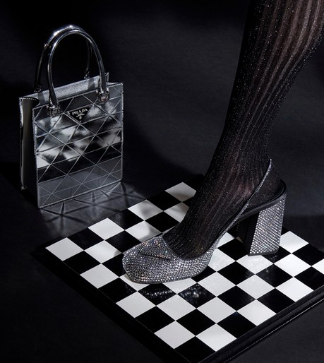 Metallic silver Prada handbag and a glittery, block-heeled shoe on a black and white checkered surface against a dark background.