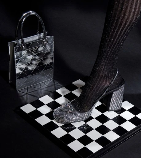 Metallic silver Prada handbag and a glittery, block-heeled shoe on a black and white checkered surface against a dark background.