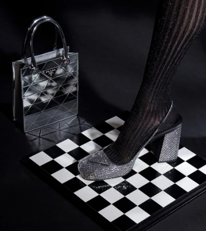 Metallic silver Prada handbag and a glittery, block-heeled shoe on a black and white checkered surface against a dark background.