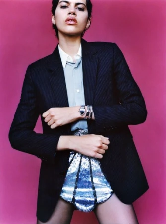 Fashionable woman posing in pinstripe blazer and silver sequin skirt against a pink background, accessorized with a silver watch and bracelets.