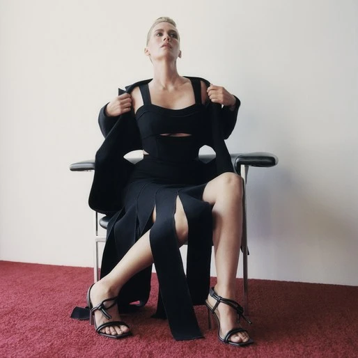 A blonde-haired woman sits in a black dress and jacket on a chair in a studio with a red carpet and white walls.