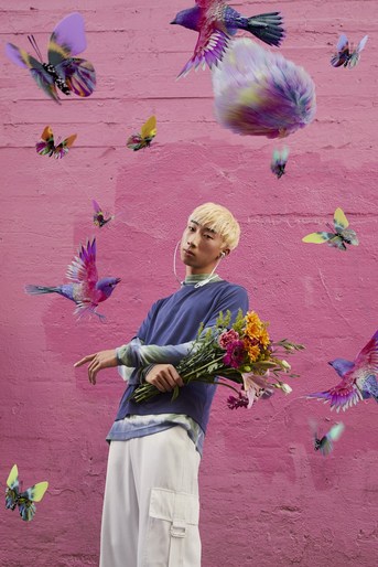 An Asian man poses outdoors against a pink wall, holding a bouquet of flowers, surrounded by digitally rendered butterflies and birds.