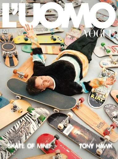 Fashion magazine cover featuring Tony Hawk lying among skateboards in black pants and color-striped sweater on a gray backdrop.