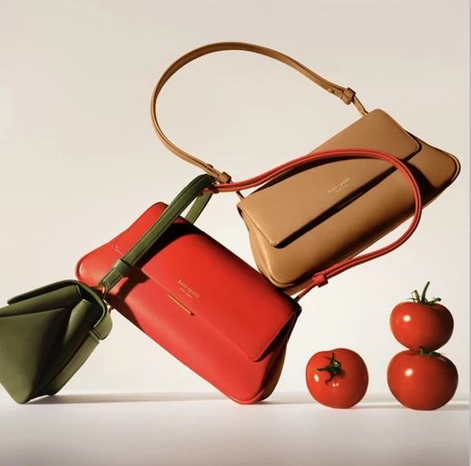 Tan, red, and green Kate Spade purses are arranged with several ripe red tomatoes on a soft white gradient background.