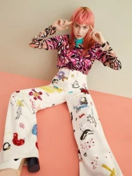 Woman with pink hair wearing patterned top and illustrated white trousers seated against pink backdrop