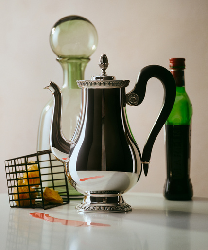 Still life shot of a silver coffee pot on a white surface, accented by a green glass bottle, beverage, and metal basket.
