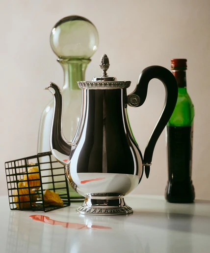 Still life shot of a silver coffee pot on a white surface, accented by a green glass bottle, beverage, and metal basket.