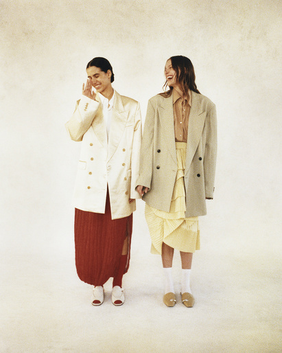 Two women in oversized blazers and skirts pose in a bright photo studio; one smiles with hand raised, the other laughs, showing teeth.