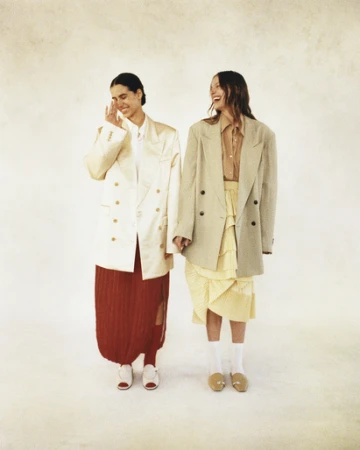 Two women in oversized blazers and skirts pose in a bright photo studio; one smiles with hand raised, the other laughs, showing teeth.
