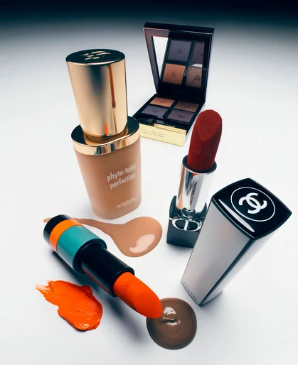 Makeup still life featuring lipstick, foundation, eyeshadow palette, and bronzer on a white surface with a gradient grey background.