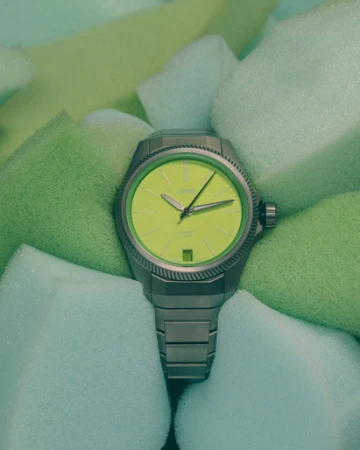 Oris watch with a stainless-steel band and a neon-yellow face nestled into a background of soft-green foam cubes, showcasing its design and color.
