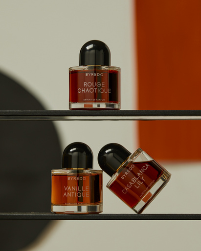 Three Byredo perfume bottles with different fragrances (Rouge Chaotique, Vanille Antique, Casablanca Lily) displayed on black shelving.