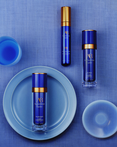 Three blue and gold Augustinus Bader skincare bottles are displayed on a blue textured background with a blue ceramic plate and cup.