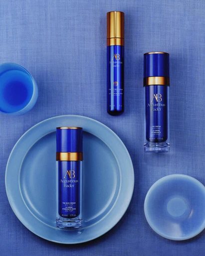 Three blue and gold Augustinus Bader skincare bottles are displayed on a blue textured background with a blue ceramic plate and cup.