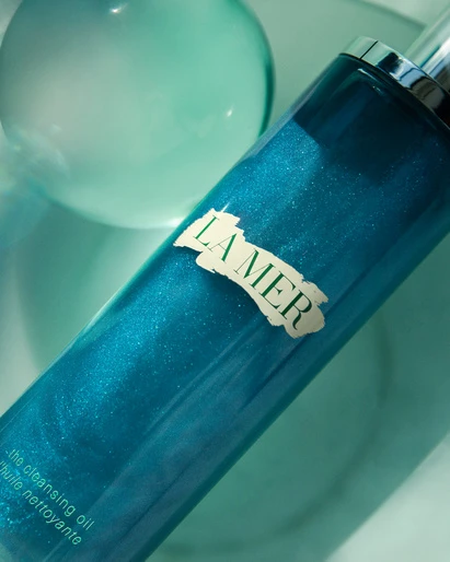 Close-up of a teal La Mer cleansing oil bottle with a spherical background, showcasing the brand's skincare line.