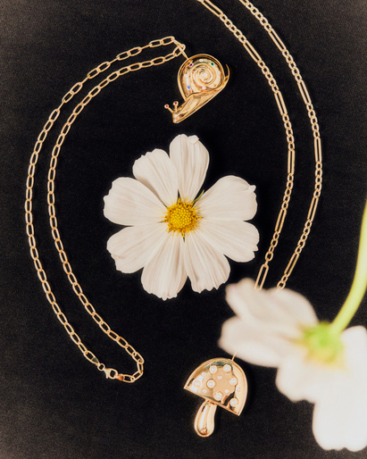 Botanical jewelry flat lay: A daisy and two necklaces with gold mushroom and snail pendants on a dark surface, studio lighting.