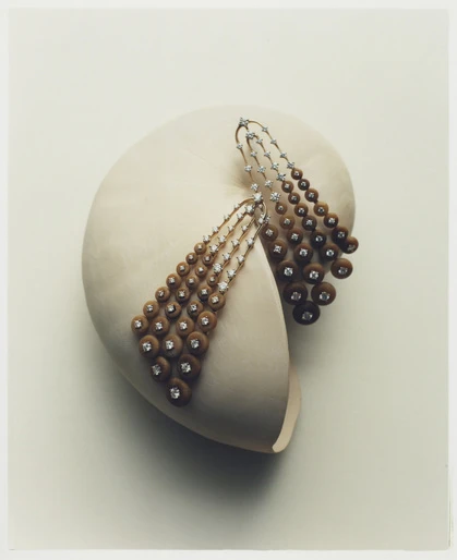Elegant dangling earrings with brown spheres and diamond accents, resting on a smooth, light-colored shell-like surface, studio lighting.