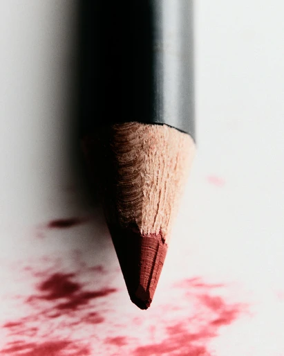 Macro shot of a red eyeliner pencil with a dark barrel, resting on white paper, with red pigment smeared on the paper surface.