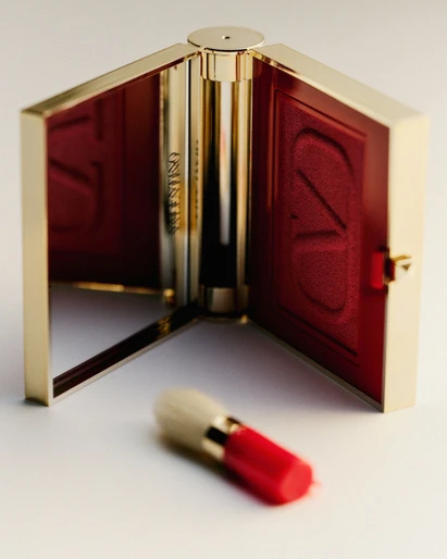 Close-up of a gold Valentino compact with red interior alongside a makeup brush on a white surface. Beauty product shot.