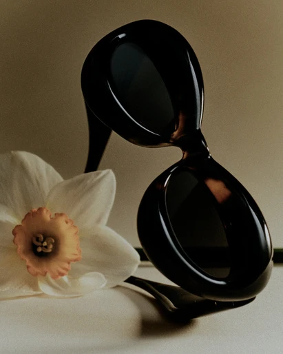 Fashion shot of tortoiseshell sunglasses next to a white daffodil against a tan background in a studio with soft lighting.
