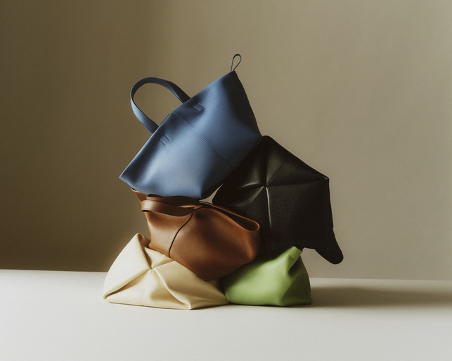 Studio shot of stacked handbags in blue, black, brown, cream, and green against a neutral background, creating a simple product display.