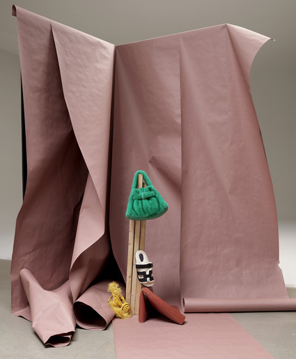 Still life shot with a green faux fur bag, patterned sandal, and orange coin purse, posed in front of a pink paper backdrop.