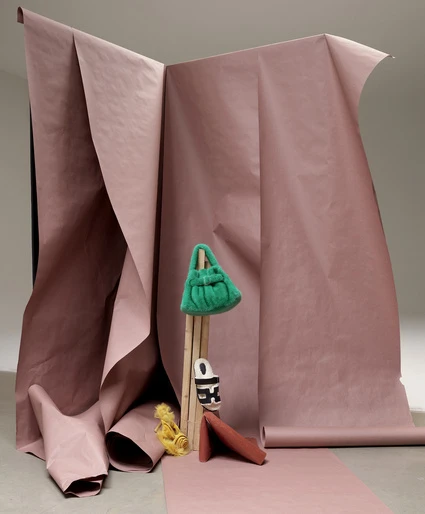 Still life shot with a green faux fur bag, patterned sandal, and orange coin purse, posed in front of a pink paper backdrop.