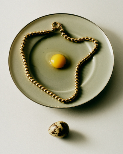 Overhead shot of gold bead necklace arranged around a raw egg yolk on a plate, and a quail egg on a white surface.