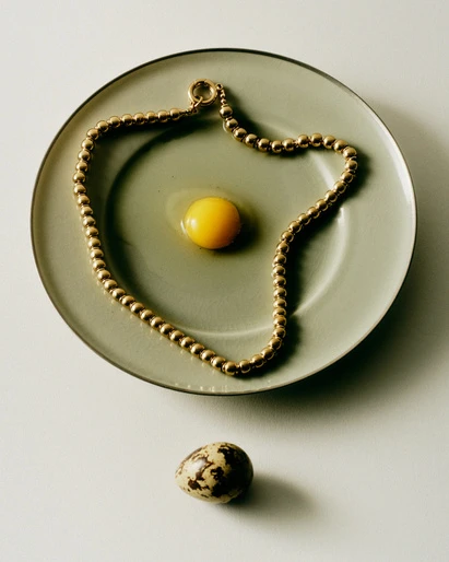 Overhead shot of gold bead necklace arranged around a raw egg yolk on a plate, and a quail egg on a white surface.