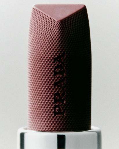 Close-up of a patterned, mauve Prada lipstick tube with a geometric top, standing on a white and black striped base.