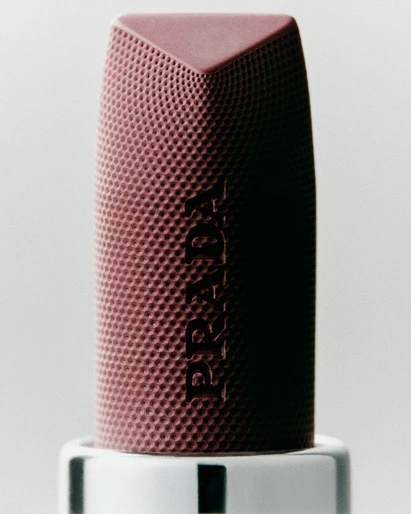 Close-up of a patterned, mauve Prada lipstick tube with a geometric top, standing on a white and black striped base.