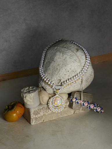 Elegant jewellery still life: pearl necklace with opal pendant on stone plinth, accented by a vintage-style bracelet and persimmon.