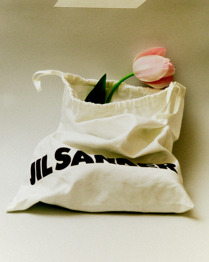 Cream-colored fabric bag with black 'JIL SANDER' text and a pink tulip with a green stem against a white background.