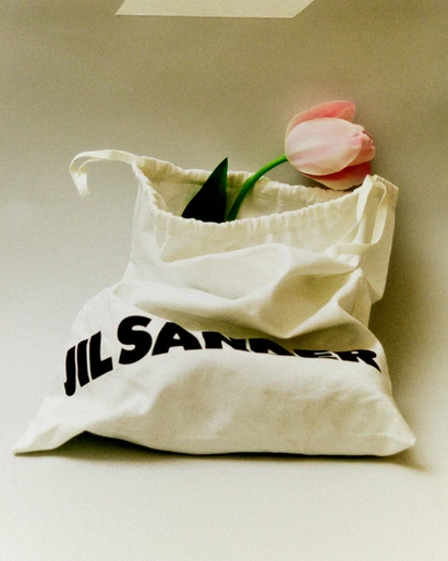 Cream-colored fabric bag with black 'JIL SANDER' text and a pink tulip with a green stem against a white background.