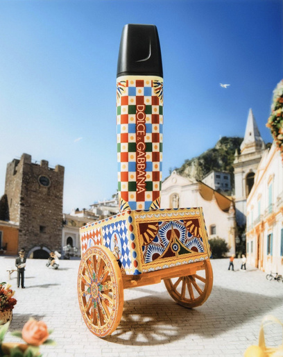 Dolce & Gabbana perfume bottle displayed on an ornate wheeled cart in an outdoor setting with buildings and blue sky.