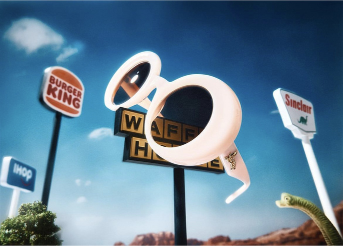 Surreal, low-angle shot of oversized sunglasses in a roadside scene with restaurant signs and a dinosaur figure against a bright blue sky.