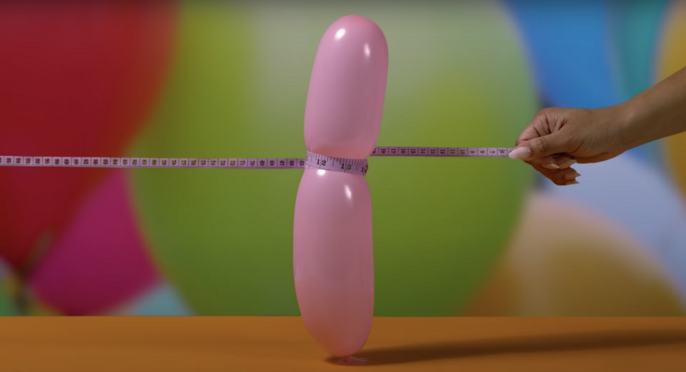 Pink balloon is measured with a pink measuring tape against a colorful blurred background and bright orange surface in a studio setting.