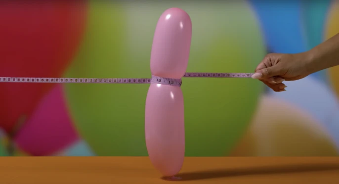 Pink balloon is measured with a pink measuring tape against a colorful blurred background and bright orange surface in a studio setting.