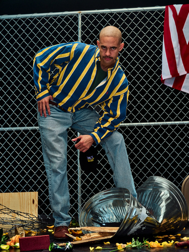 Man in blue and yellow striped jacket poses near a chain-link fence with crushed metal, creating an edgy and unconventional composition.