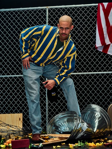Man in blue and yellow striped jacket poses near a chain-link fence with crushed metal, creating an edgy and unconventional composition.