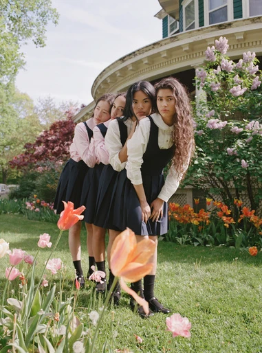 Five young women in school uniforms stand in a line outdoors among colorful flowers, with a house visible in the background.