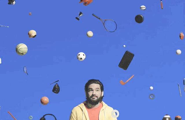 Man looking up at an assortment of floating sports equipment, a phone, a trophy, and headphones on a blue background.