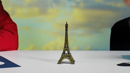Eiffel Tower miniature stands on a white table in front of a cloudscape backdrop between two people in red and black shirts.