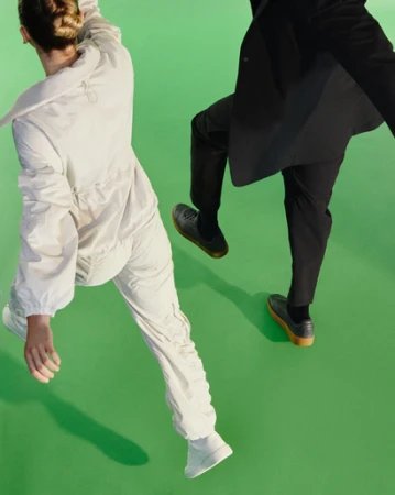 Two people in motion against a bright green backdrop, one wearing white athletic wear and the other dark trousers and a jacket.
